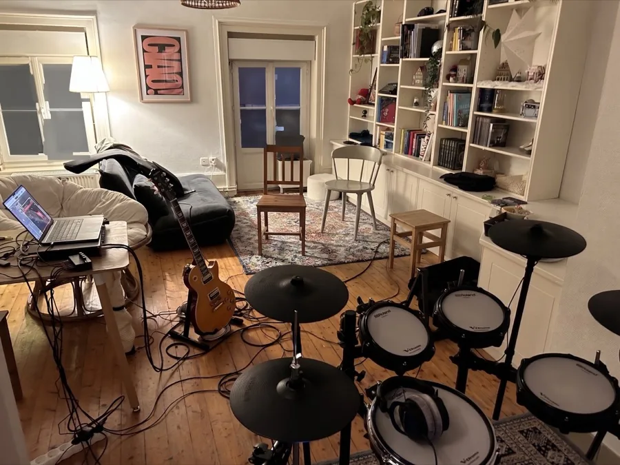 Le séjour aménagé en studio : batterie électronique au premier plan, guitare électrique sur pied, ordinateur de régie sur une table.